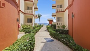 A corridor with a pathway flanked by neatly trimmed hedges, leading to a building with balconies and a view of the sea.
