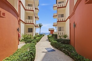 Ein Gang mit einem Weg, der von ordentlich beschnittenen Hecken gesäumt wird, führt zu einem Gebäude mit Balkonen und Blick auf das Meer.