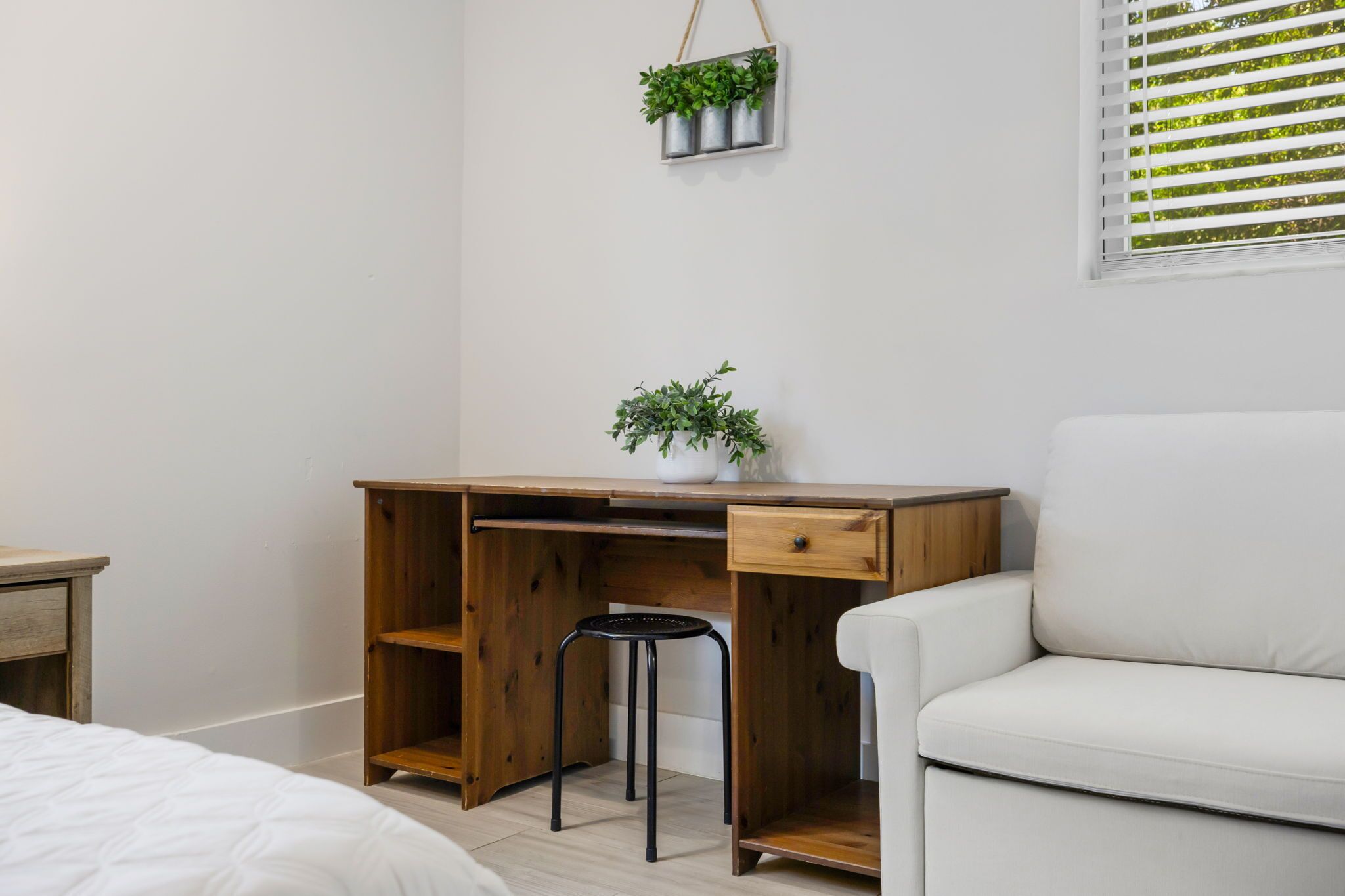 A bedroom with a wooden desk, a white chair, a bed, and a hanging plant.