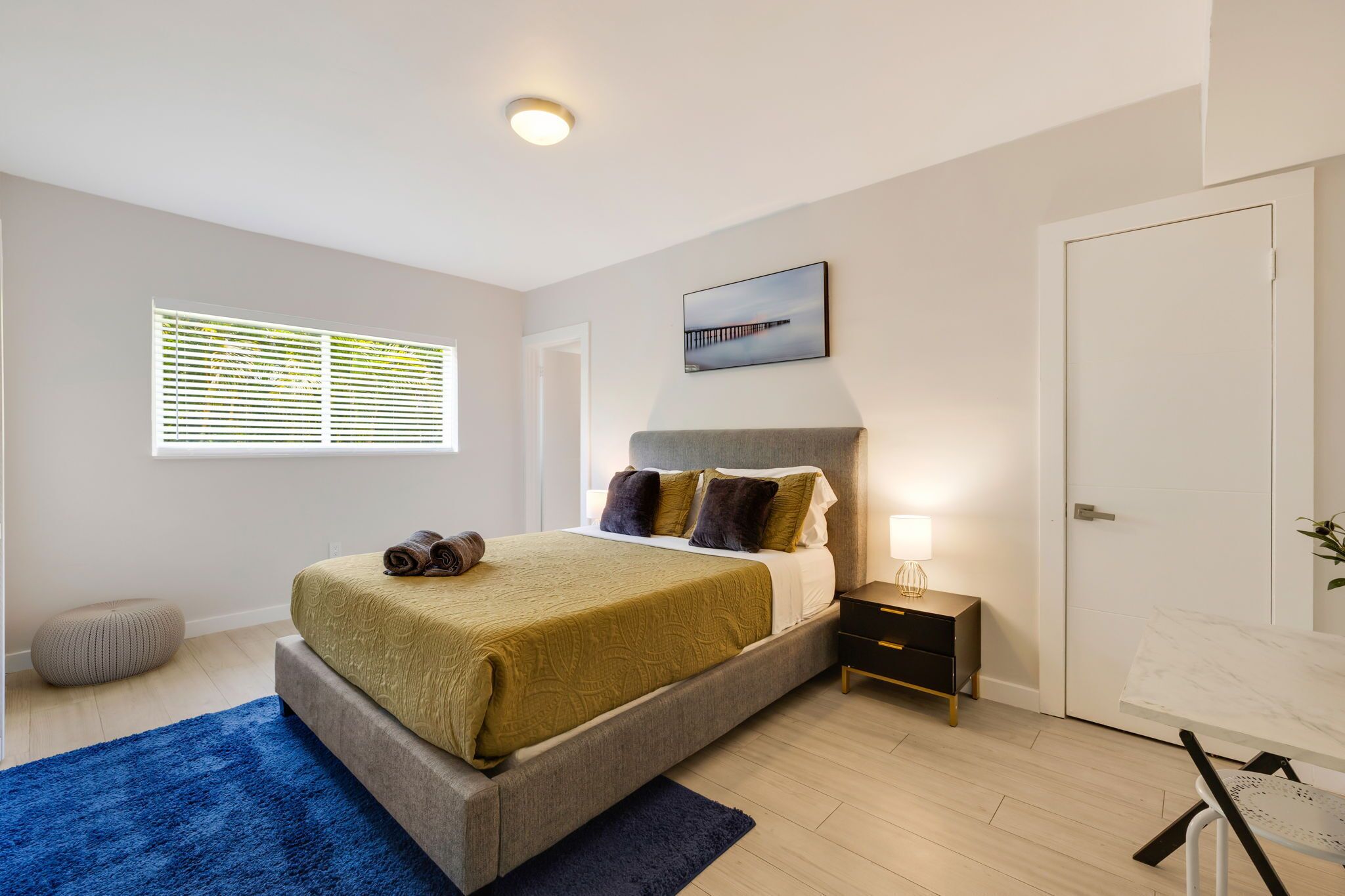 A bedroom with a bed, bedside table, lamp, window with blinds, and a door.