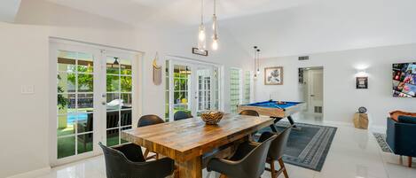 A spacious room with a wooden dining table, black chairs, a pool table, and a pool visible through glass doors.