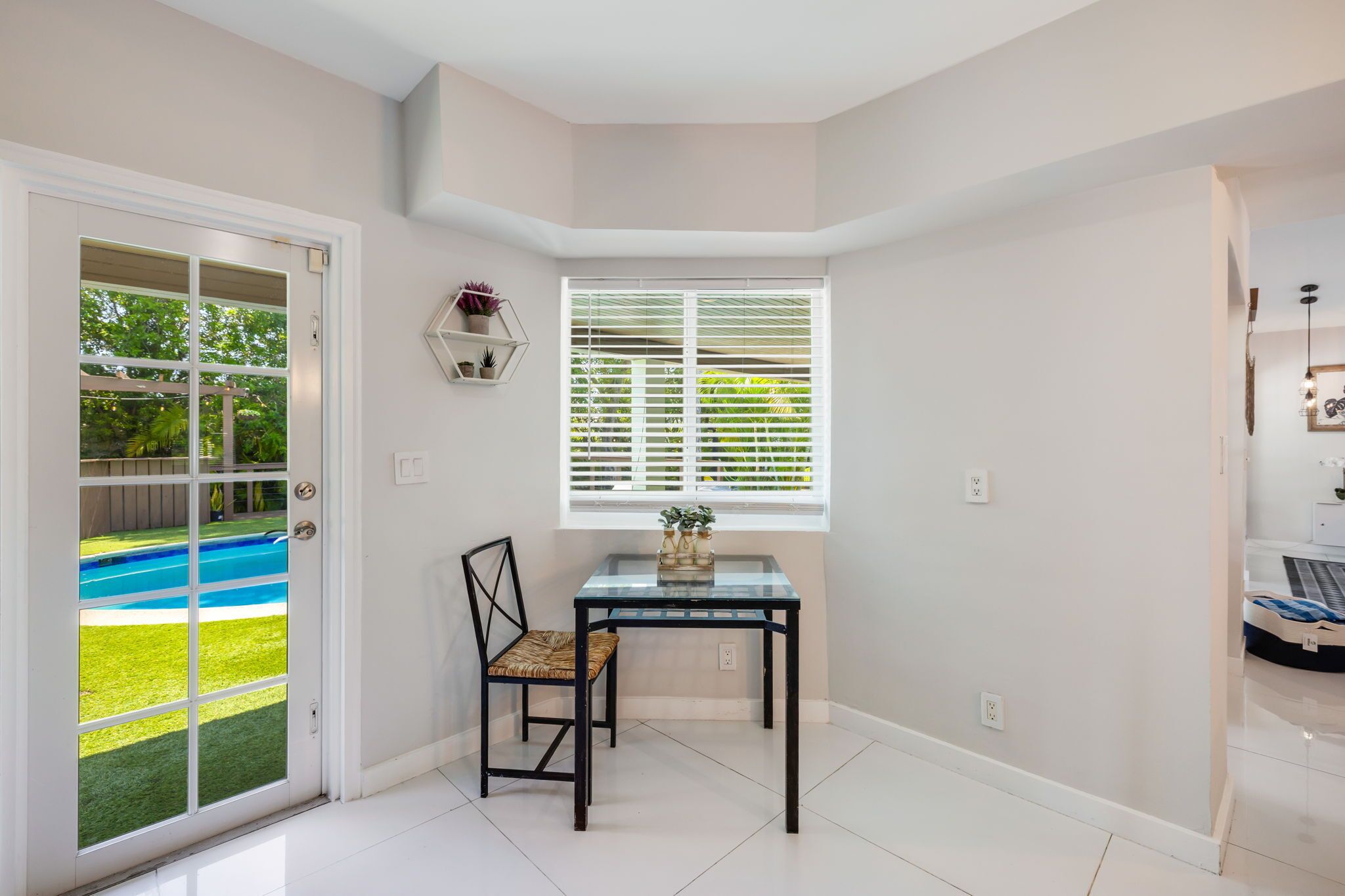 A room with a glass table, a chair, a window with blinds, a door leading to a pool, and a small shelf with plants.