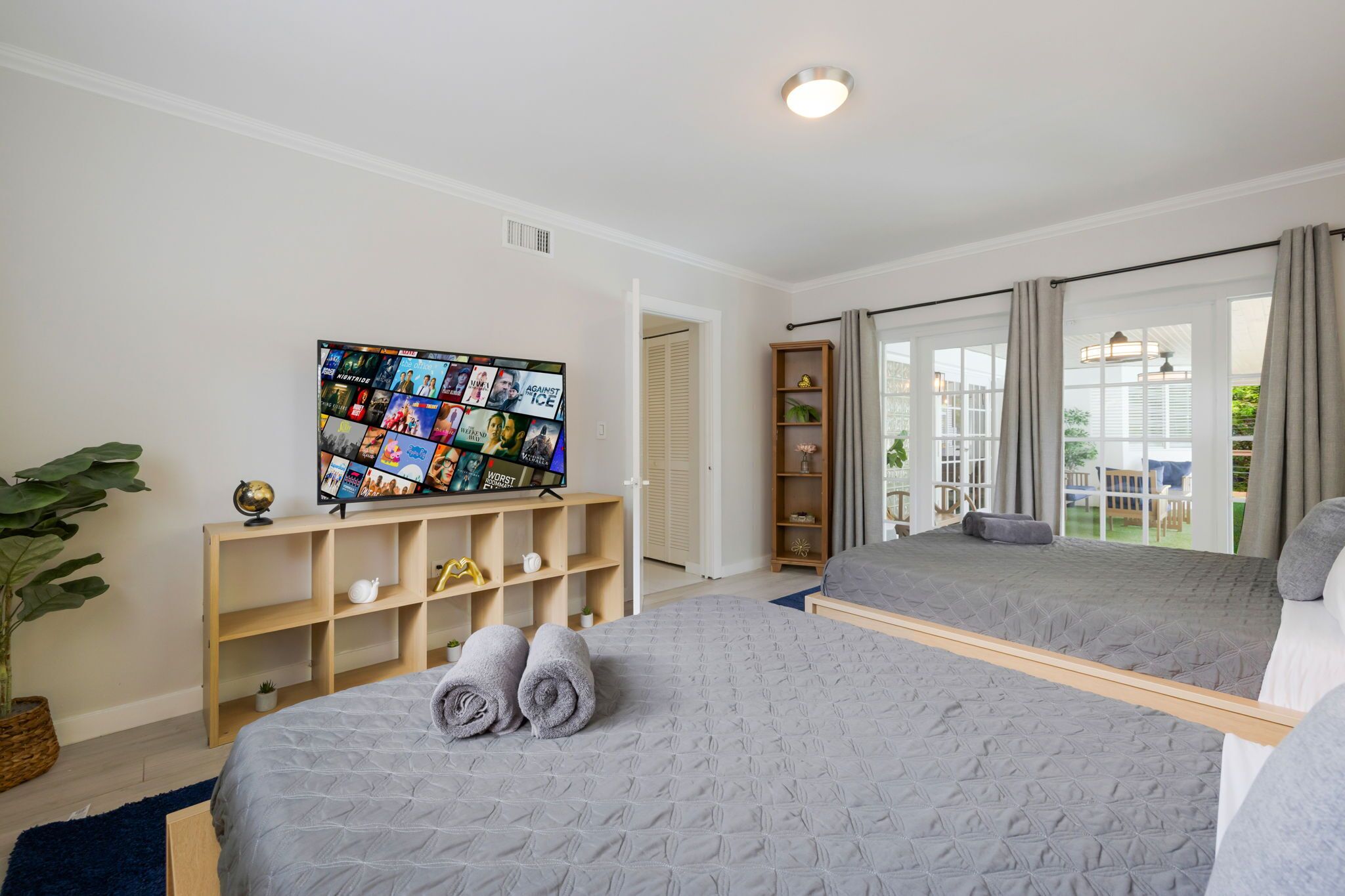 A hotel room with two beds, a television displaying travel photos, a bookshelf, and a large window with curtains.