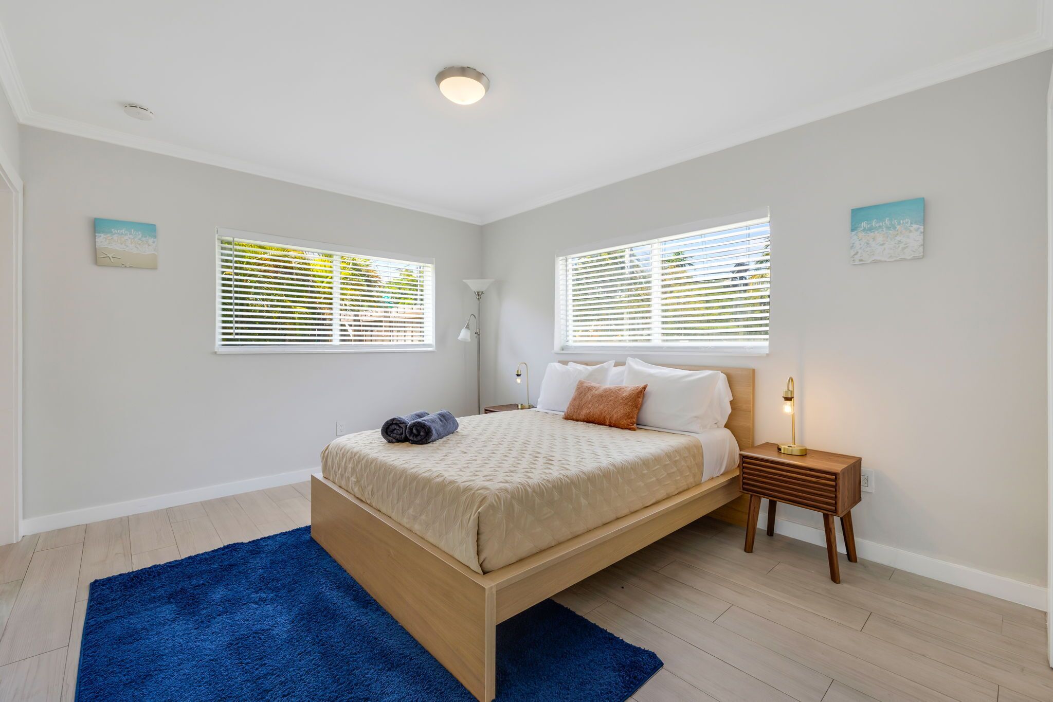 Un dormitorio con cama, dos ventanas con persianas, una lámpara de noche y una alfombra azul.