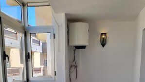 Ein Zimmer mit einem hellen Sofa, einem Fenster mit Blick auf Gebäude, einer Wandleuchte und einem Deckenventilator.