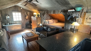 A rustic cabin interior with a wooden floor, a sofa, a coffee table, a dining table, a kitchen area, and a ceiling fan.