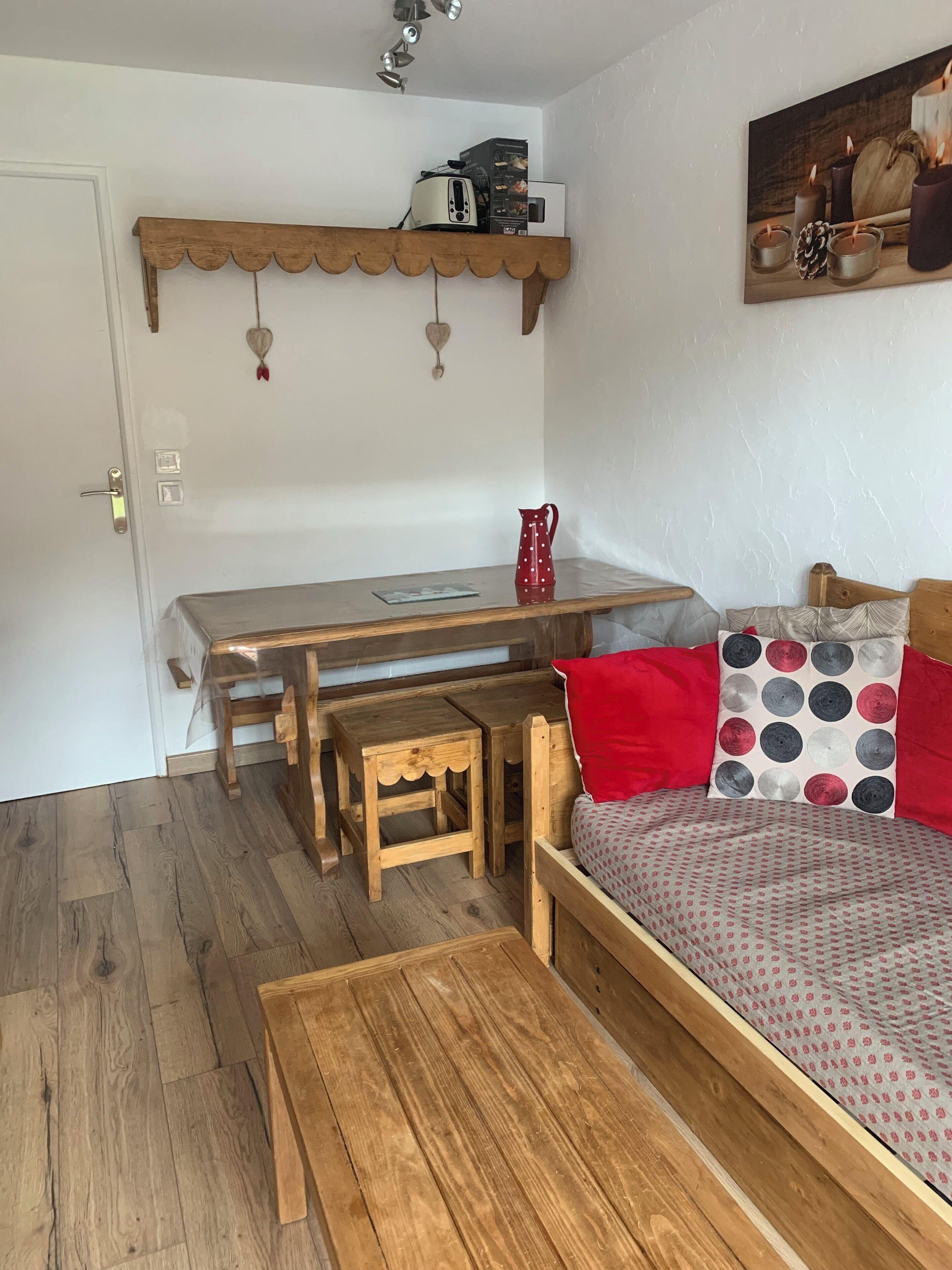 A cozy room with a wooden bed, a small wooden table, a wooden desk, and a shelf with a toaster and other items.