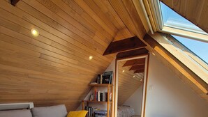 A cozy attic bedroom with a wooden ceiling, a built-in bookshelf, a gray sofa with yellow cushions, and a skylight.