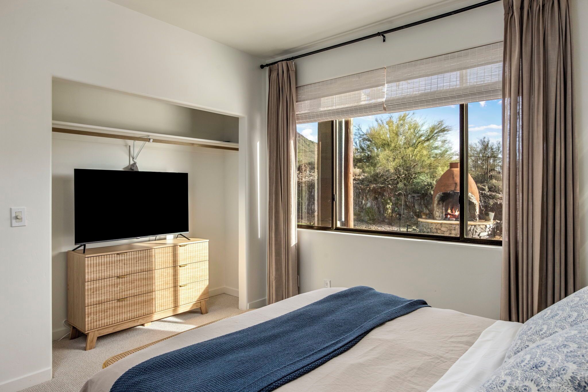 Une chambre à coucher avec un lit, un téléviseur posé sur un meuble en bois et une fenêtre donnant sur un immeuble et des arbres.
