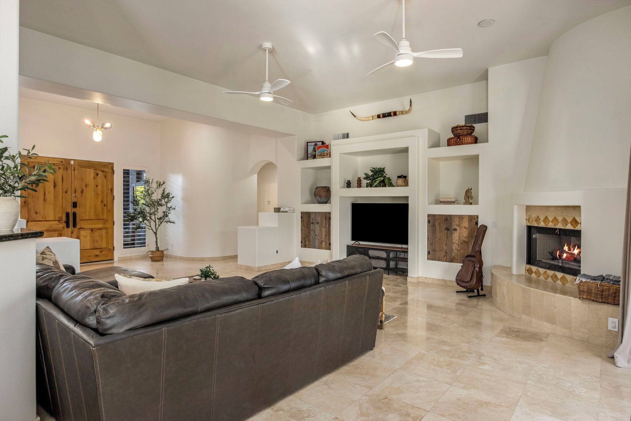 A spacious living room with a dark leather sofa, a flat-screen TV, and a fireplace. There are ceiling fans and shelves with decorative items.