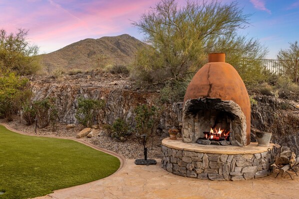 A stone-built outdoor fireplace with a fire, surrounded by a landscaped garden and a mountainous backdrop.
