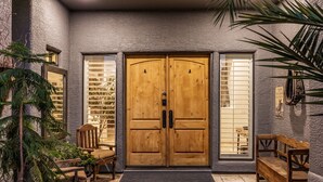 Une porte à deux battants en bois, dotée d’une poignée noire, flanquée de deux fenêtres équipées de stores, d’un banc et d’un pot de fleurs.