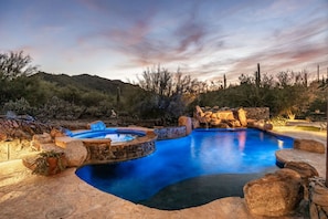 A swimming pool with a stone border and a waterfall feature, surrounded by desert vegetation and a mountainous backdrop.