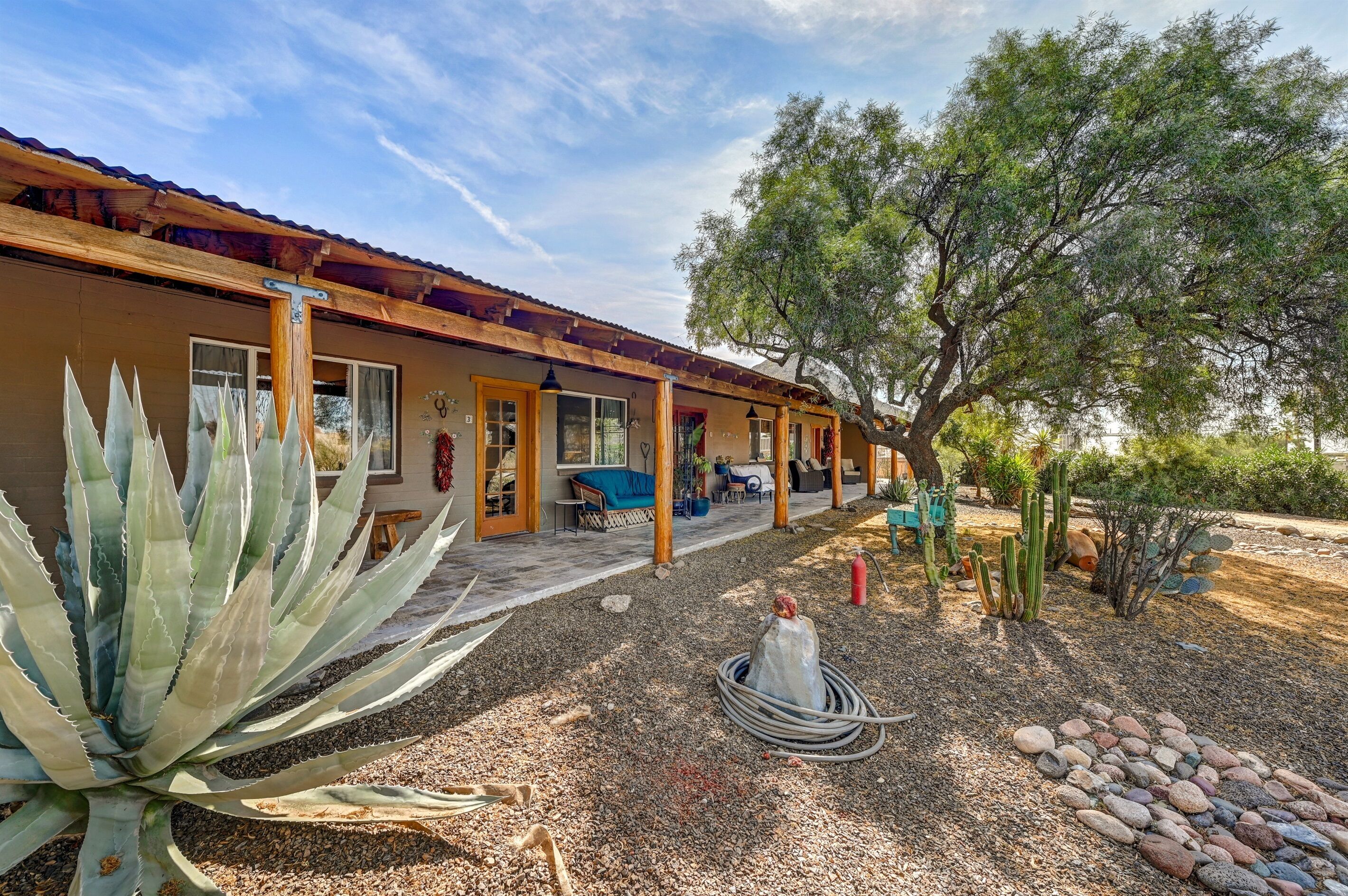 Ein Haus mit Holzfassade, einer überdachten Terrasse und einem Wüstenpflanzen-Garten mit Kakteen und einer Agave.