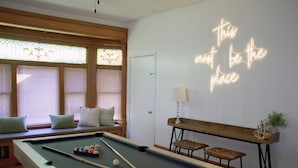 Een pooltafel met biljartballen, een neonbord, een bank en een raam met verstelbare luiken.