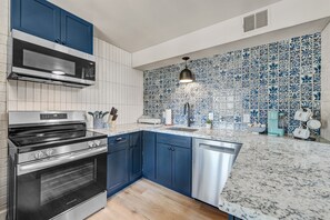 A kitchen with blue cabinets, a stainless steel oven, a microwave, and a sink with a blue patterned backsplash.