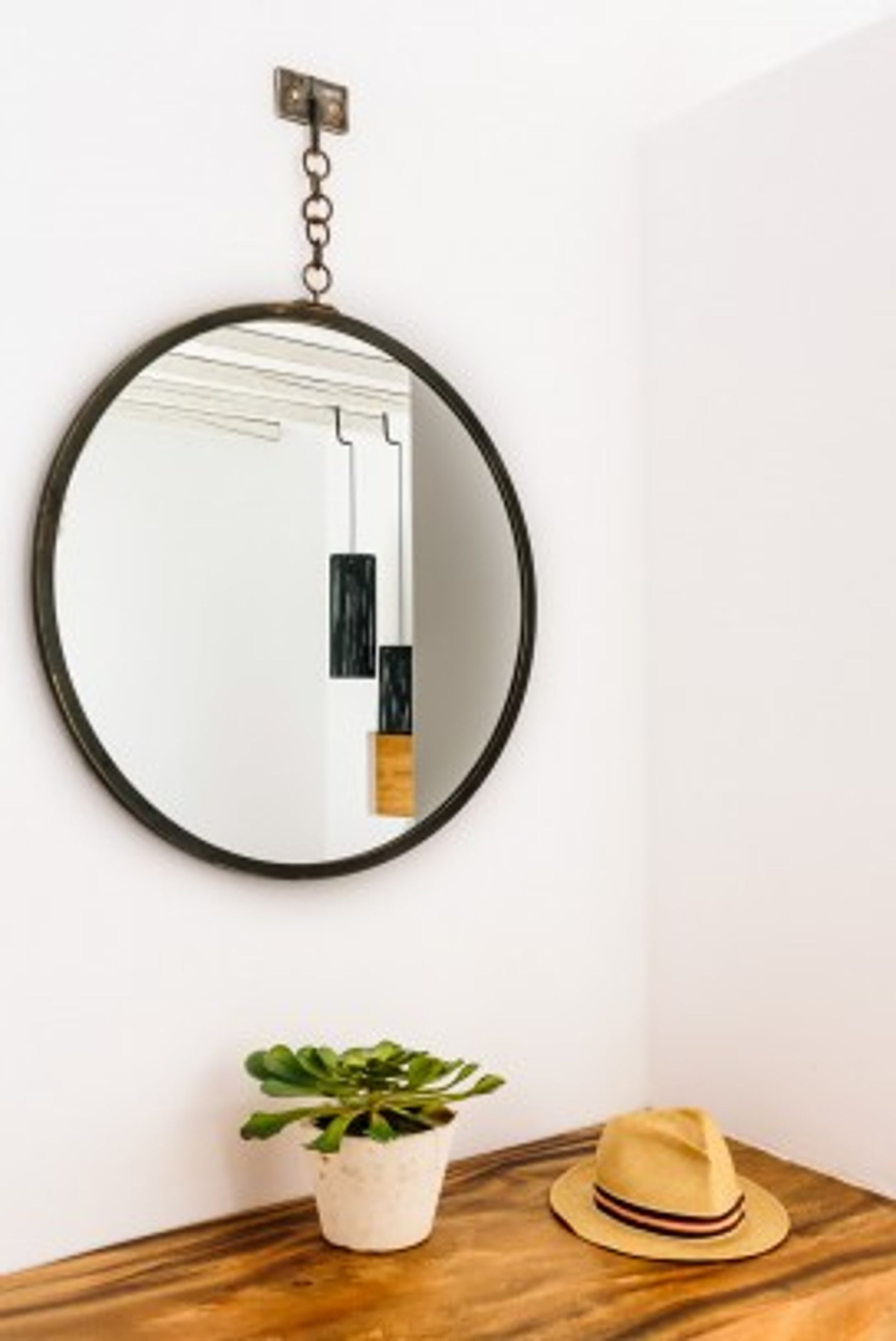 A round mirror with a black frame hanging on a white wall. Below the mirror, there is a small potted plant and a hat on a wooden surface.