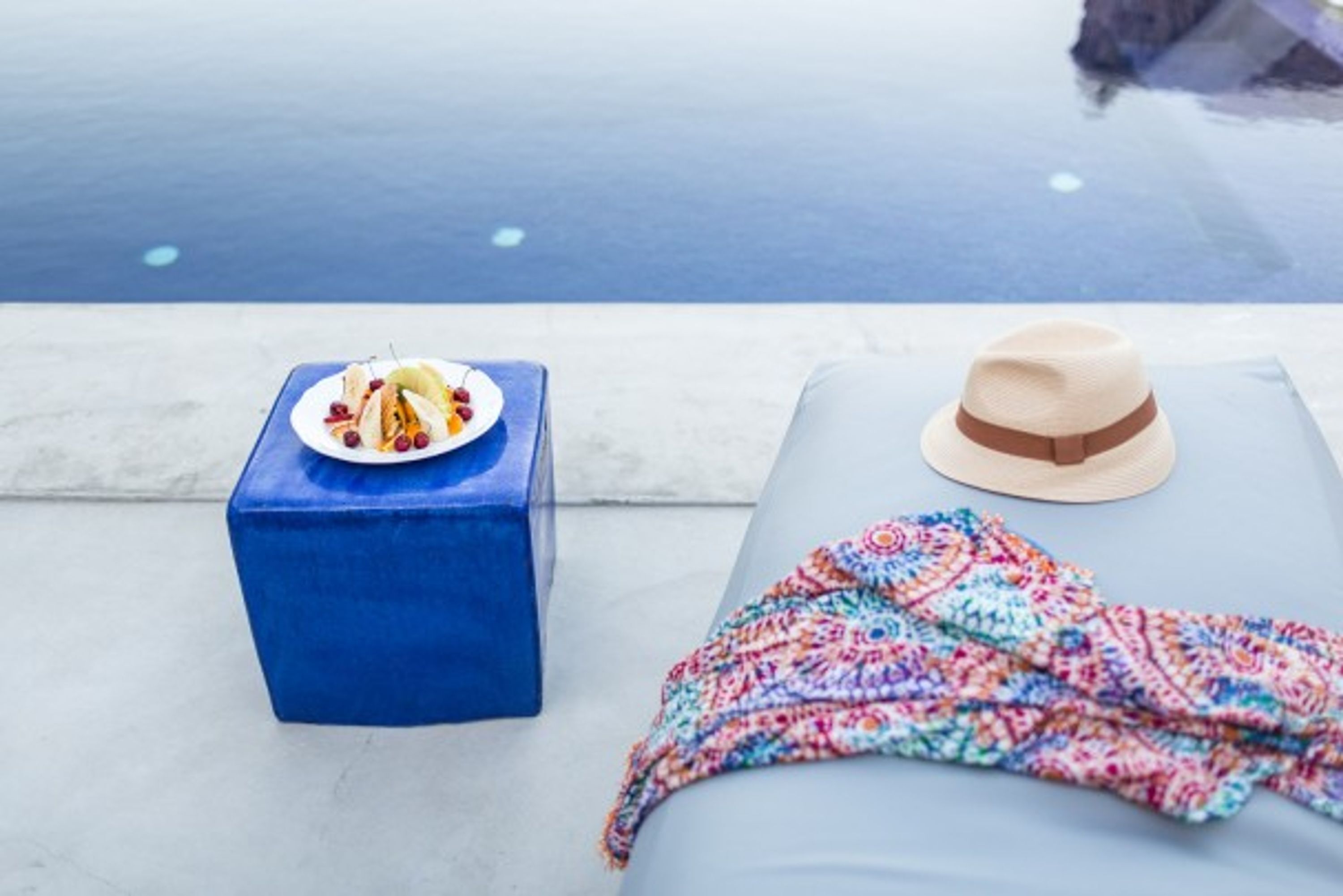 Escena junto a la piscina con un taburete azul, un sombrero y una manta colorida.