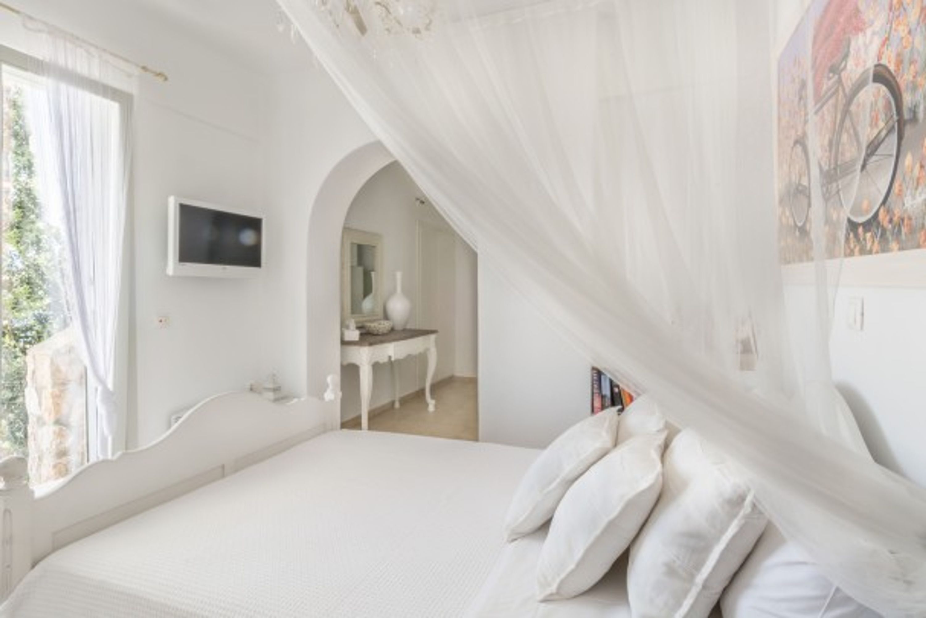 A bedroom with a bed, a TV, a window with curtains, a bedside table, and a painting on the wall.