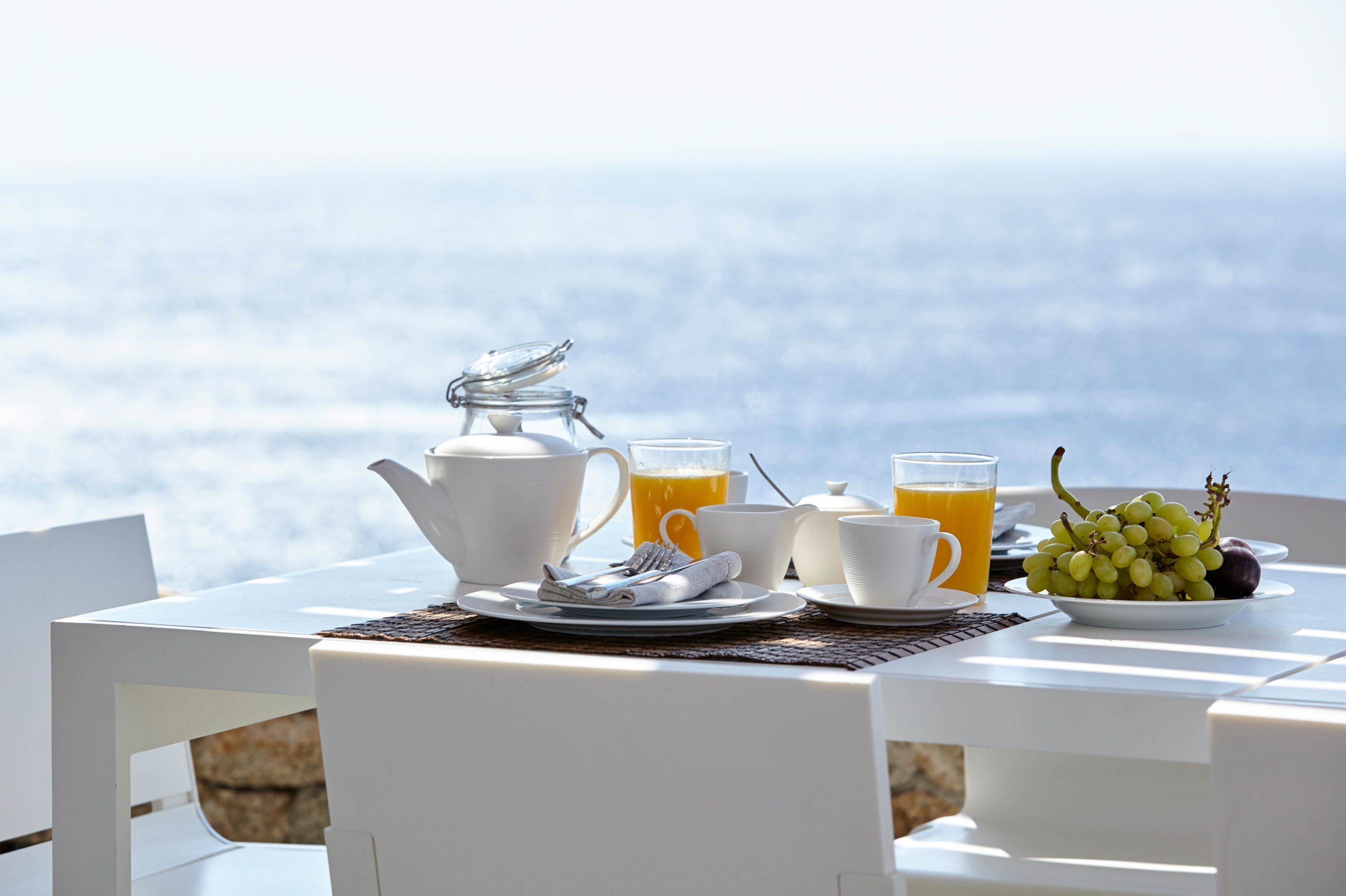 Een ontbijt opstelling met een theepot, kopjes en een glas sinaasappelsap op een tafel met uitzicht op de oceaan.