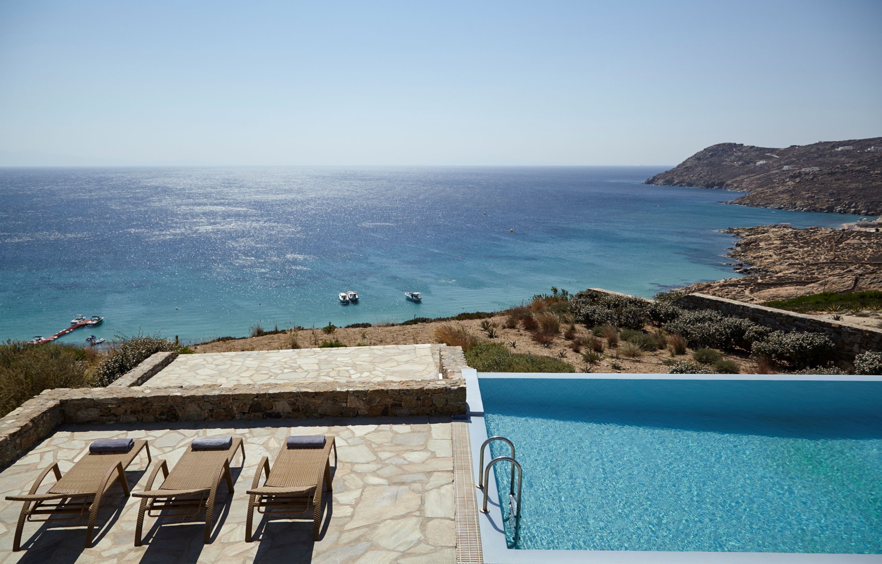 A pool with a view of the sea, lounge chairs, and rocky terrain.