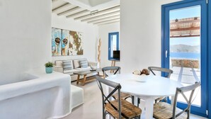A modern living room with a white sofa, a round table, and chairs. There is a painting on the wall and a window with a view of the sea. - 2 Bedroom Villa with private pool (Panormos)