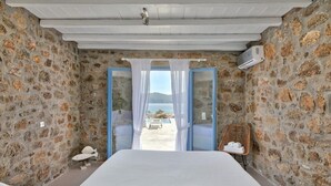 A bedroom with stone walls, a bed, a view of the sea, and a wicker chair. - 2 Bedroom Villa with private pool (Panormos)