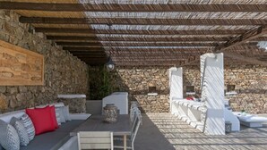 Um pátio externo com paredes de pedra, teto de madeira e área de estar com almofadas e uma mesa.