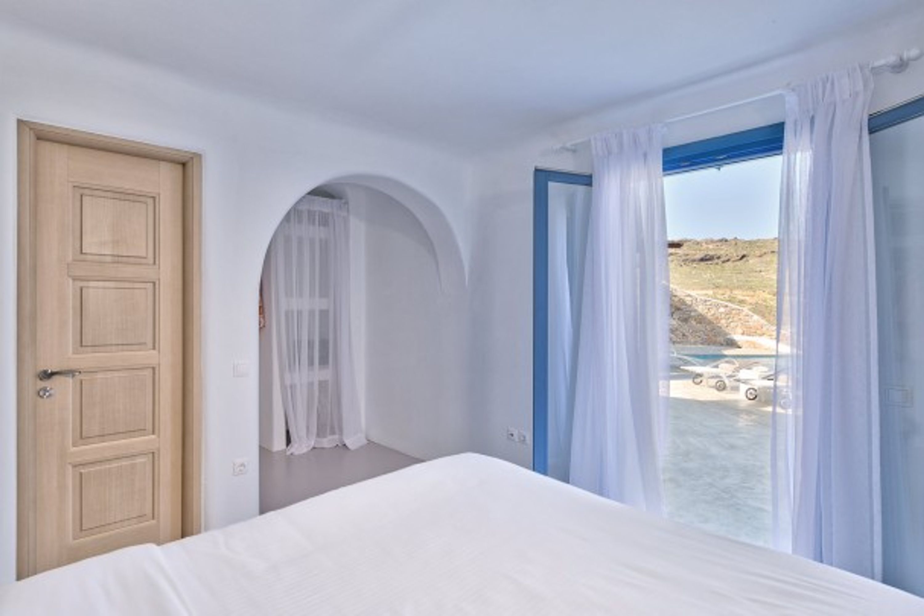 Een slaapkamer met een bed, een houten deur, een boogvormige deuropening en een raam met witte gordijnen.
