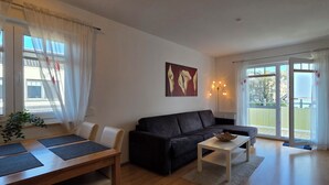 A living room with a sofa, coffee table, dining table with placemats, a painting, and a view through a glass door. - Beachfront, 2-room apartment on the 1st floor with WLAN (Graal-Müritz)
