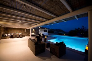 An outdoor pool area with wicker furniture, a stone wall, and a view of the pool at dusk. - Superior villa Divine/private pool, sea view (Kalafatis)