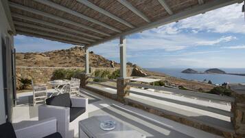 Un espacio de estar al aire libre techado con mobiliario de mimbre blanco y vistas al mar y a las colinas.