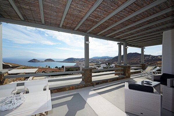 Un espace extérieur aménagé avec des meubles blancs, abrité et offrant une vue sur la mer et les collines lointaines.
