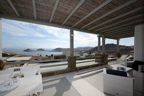 Un espace extérieur aménagé, abrité et doté de mobilier blanc, offrant une vue sur un paysage côtier avec des collines et une étendue d’eau.