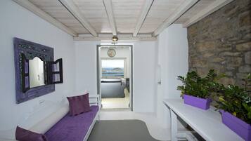 Una habitación con techo blanco, pared de piedra, cama con vistas, un banco morado y dos macetas moradas.