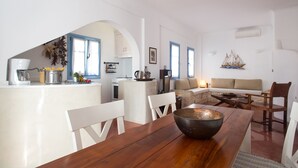 A modern kitchen with white cabinetry, a dining area with a wooden table and chairs, and a living area with a sofa and a small ship decoration on the wall. - Daphne - Two Bedroom Villa, private pool, sea view (Agia Paraskevi)