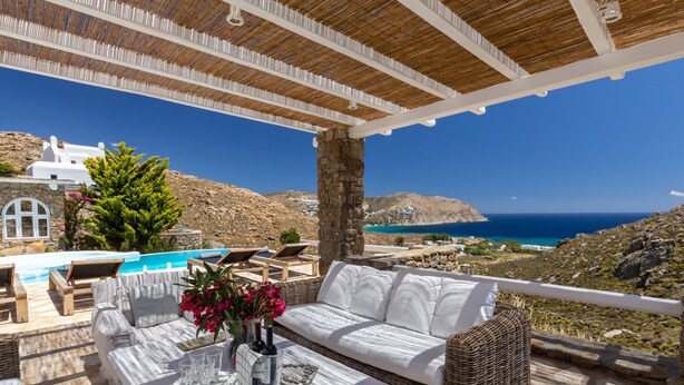 A terrace with a white sofa, wicker furniture, a table with drinks, and a view of the sea and mountains. - Rocky Mansion Mykonos (Mykonos)