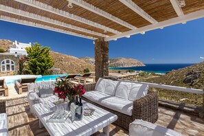 A terrace with a white sofa, wicker furniture, a table with drinks, and a view of the sea and mountains. - Rocky Mansion Mykonos (Mykonos)