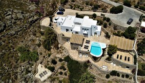 Luftaufnahme einer modernen Villa mit Schwimmbad, mehreren Autos und einer felsigen Landschaft.
