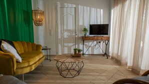 Una sala de estar con un sofá amarillo, una mesa redonda para café, un televisor sobre un mueble de madera y una puerta con cortina verde.