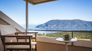 Un balcón con una silla de madera y una mesita, con vistas a un paisaje montañoso y una masa de agua.