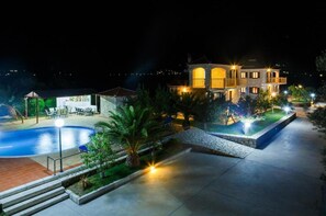 A well-lit, multi-story building with a swimming pool, outdoor seating, and landscaped gardens. - Glyfada    Villas II (Paralio Astros)