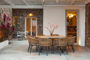 Une terrasse aux murs de pierre, avec une table ronde et des chaises en osier, une porte blanche et une porte vitrée donnant sur l’intérieur.