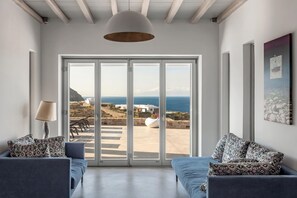 A modern living room with a large sliding glass door leading to a terrace with a view of the sea. - Artemis | 7BD minimal villa, Private pool, Jacuzzi (Mykonos)