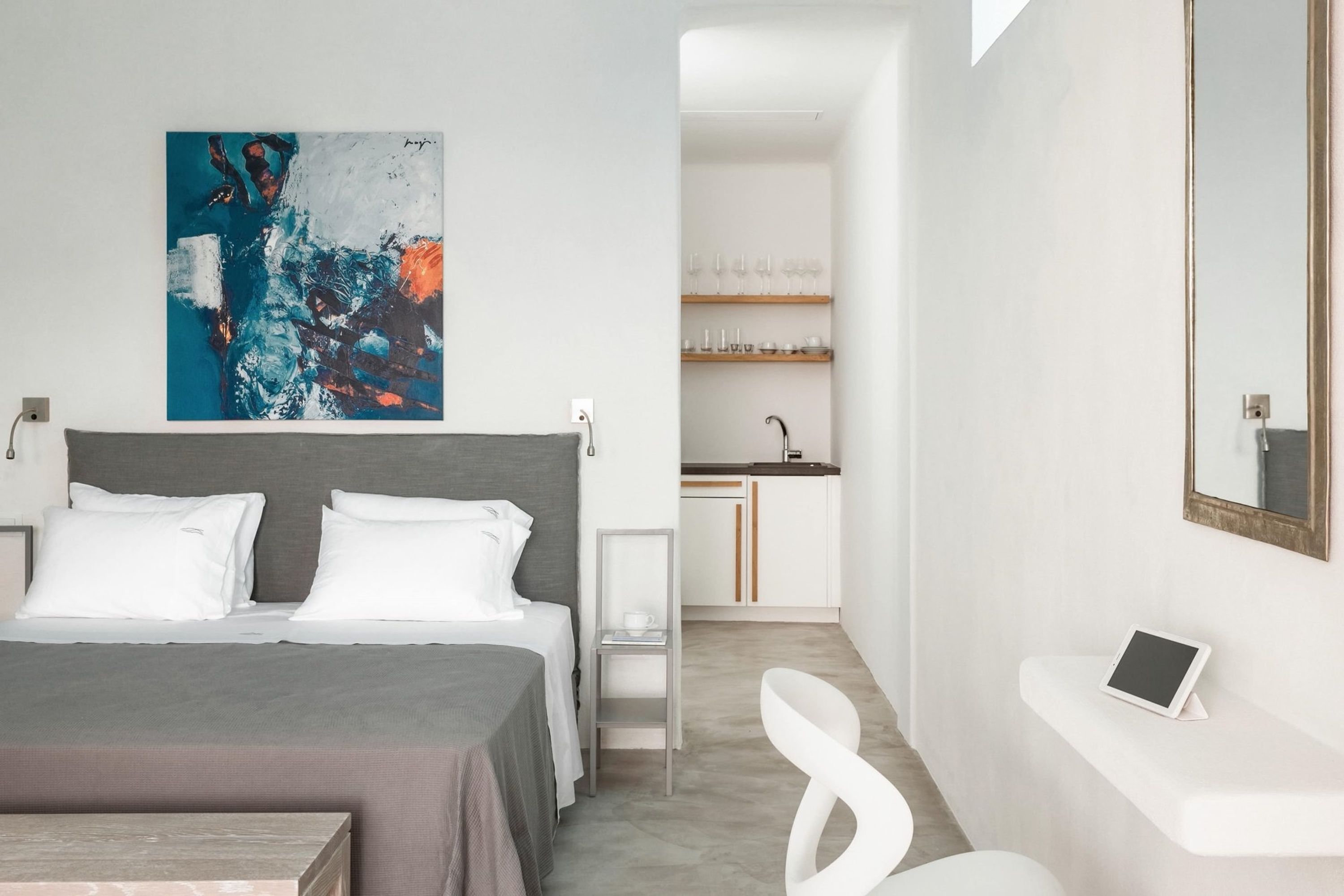 A modern hotel room with a bed, a desk, a chair, a tablet, and a painting on the wall.