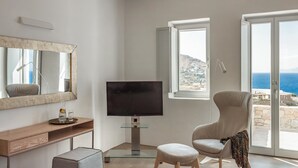 Ein modernes Wohnzimmer mit einem Flachbildfernseher, einem bequemen Sessel, einem Glastisch und einem großen Fenster mit Meerblick.