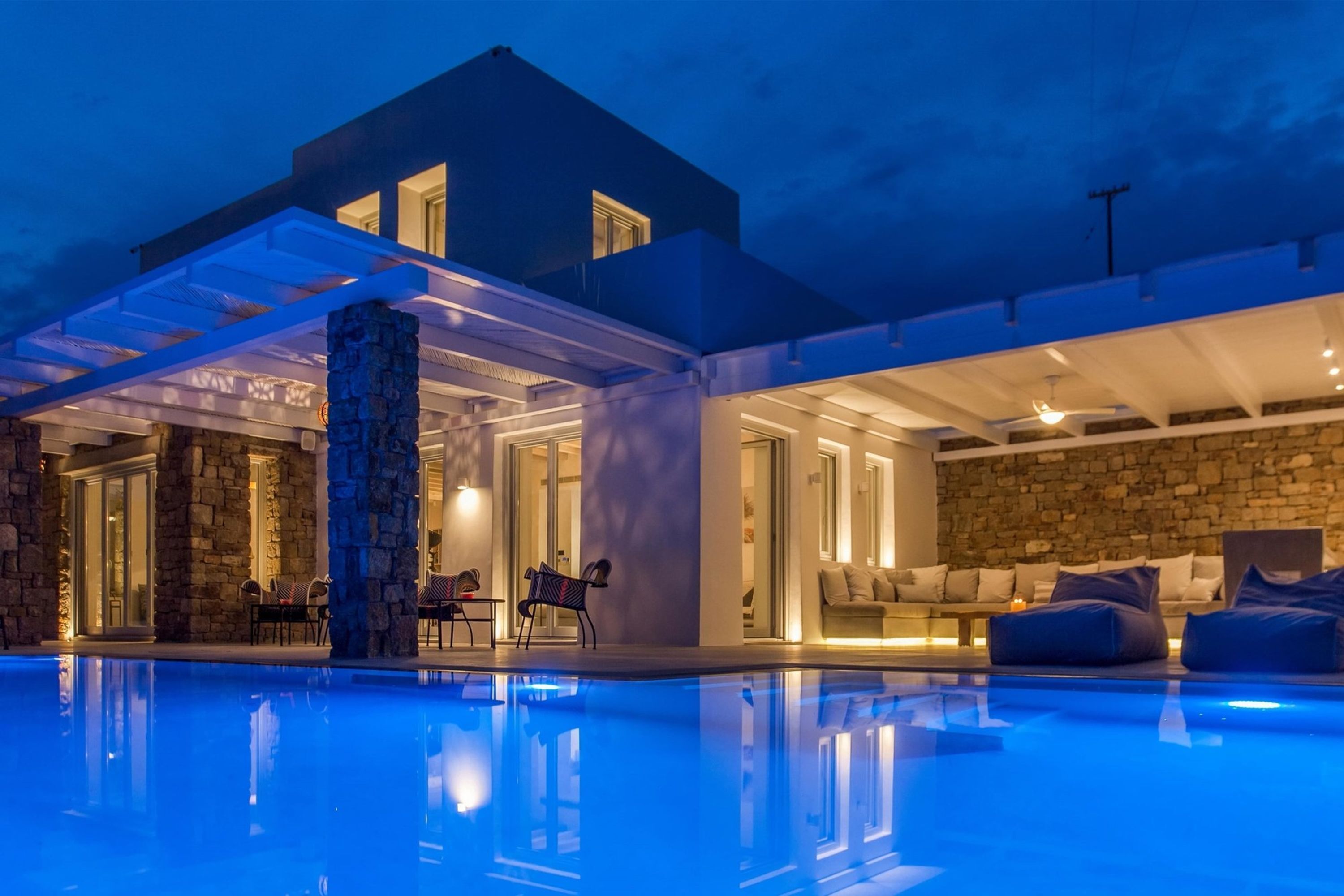 Un espace moderne aménagé au bord de la piscine, avec une piscine à eau claire, des piliers en pierre et un espace intérieur bien éclairé, doté de sièges.