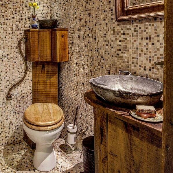 Een badkamer met een houten toilet, een grote metalen wasbak en een betegelde muur met mozaïek.