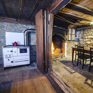 Ein rustikaler Kamin aus Stein mit Feuer, ein weißer Herd und ein Holztisch mit Stühlen in einem Raum mit Steinwänden.