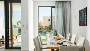 A dining area with a table set for six, a view of a pool and buildings, and a door leading to an outdoor seating area. - Opulent VILLA K on Crete (Galatas)